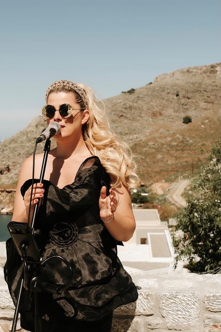 Jeris singing at a microphone with a Greek hillside behind